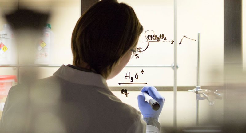 A scientist facing away from the camera, writing an equation on a whiteboard